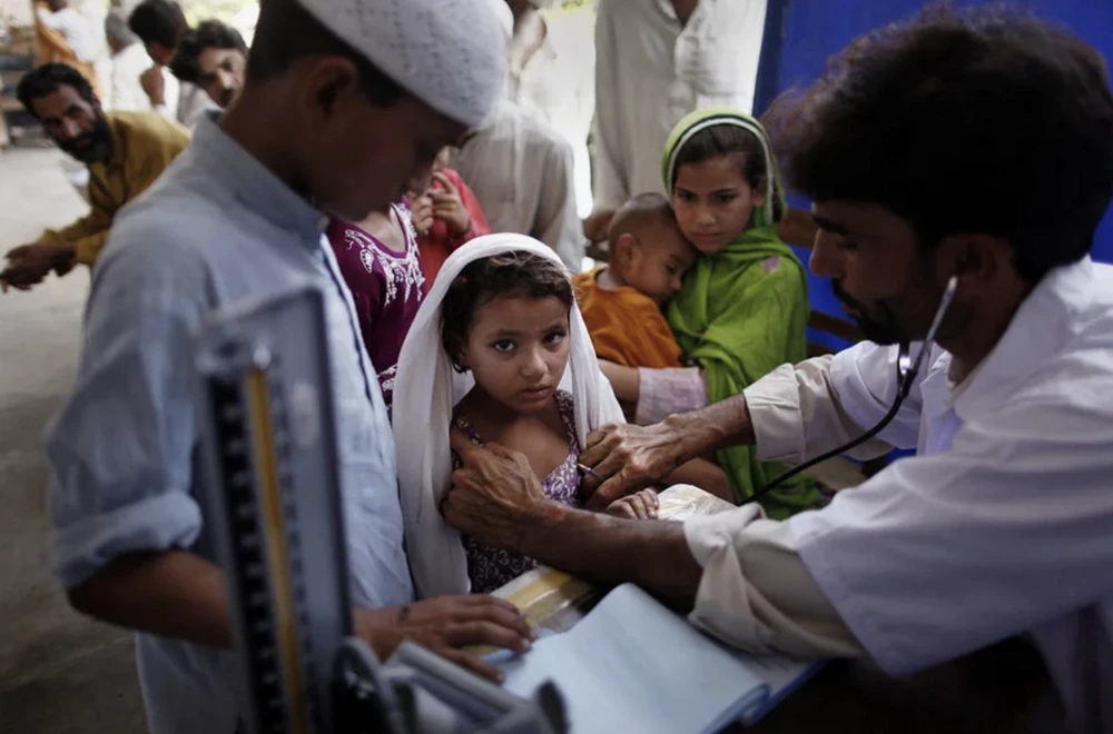 A doctor examines a young girl with a stethoscope at a crowded clinic. Other children and adults, some seated and some waiting in line, are visible in the background. The scene suggests a medical outreach or health camp.