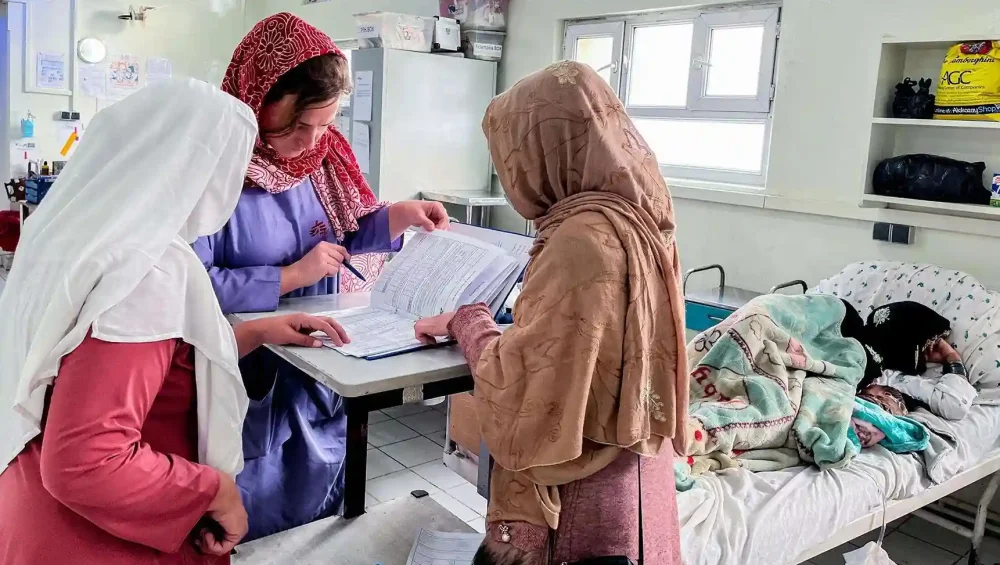 Three women in headscarves review medical documents at a table in a hospital room, while another woman lies in a hospital bed covered with blankets in the background. The setting appears bright and clinical.