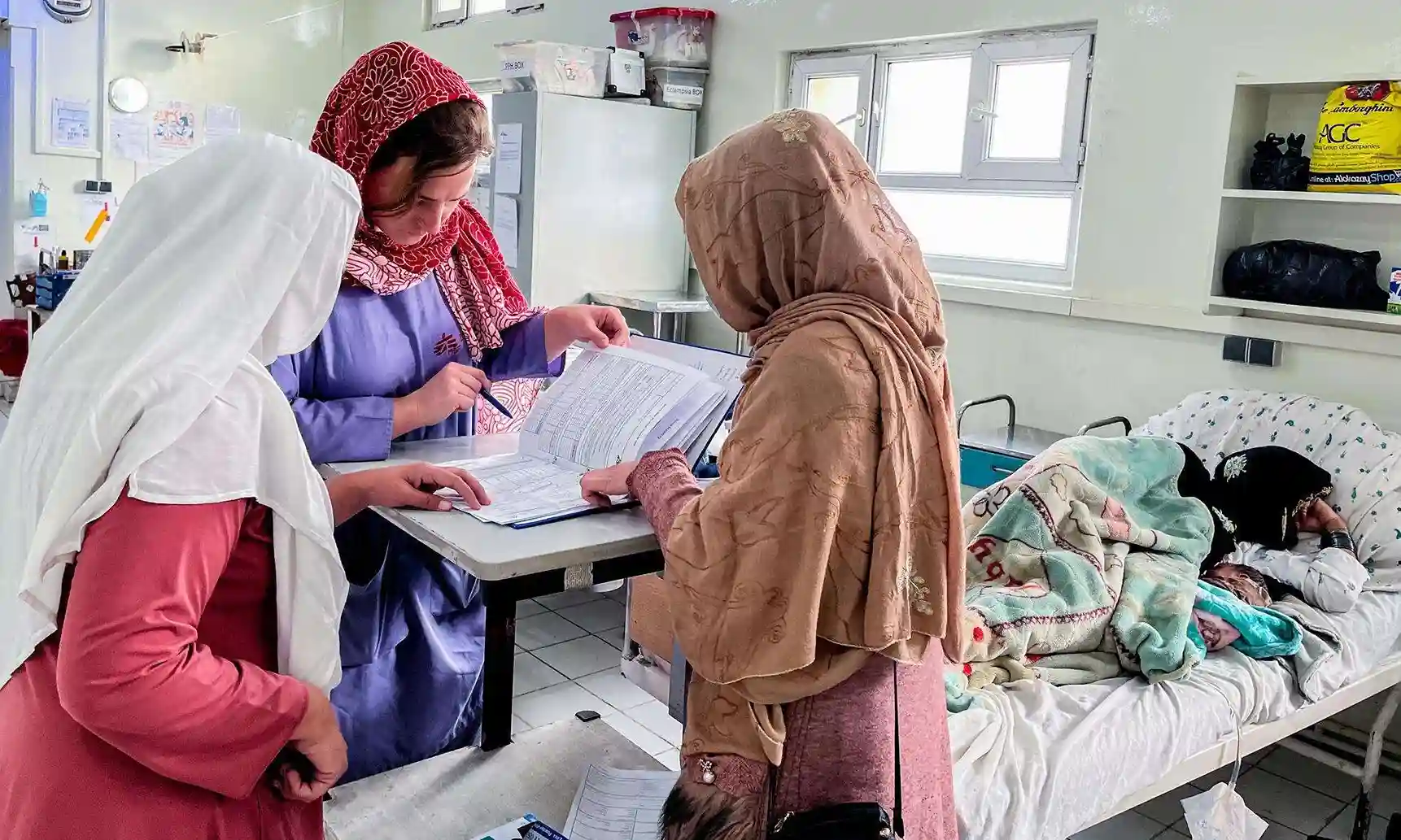 Three women in headscarves review medical documents at a table in a hospital room, while another woman lies in a hospital bed covered with blankets in the background. The setting appears bright and clinical.